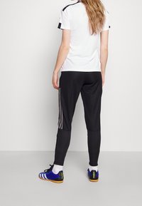 Bílá košile s krátkým rukávem a černými akcenty, doplněná černými zužujícími se kalhotami s šedými bočními pruhy a modrými sportovními botami.