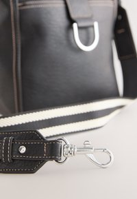 Bandoulière de sac en cuir noir avec fermoir argenté et couture beige, attachée à un sac noir avec une bandoulière rayée blanche et noire contrastante.