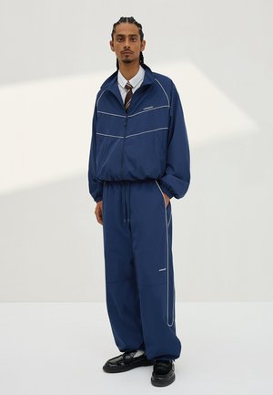 Fonott hajú férfi tengerészkék színű, fehér csíkozású melegítőben, fehér ingben, csíkos nyakkendőben és fekete cipőben, egyszerű háttér előtt.