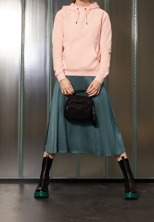 Personne portant un sweat à capuche rose, une jupe midi bleu sarcelle, des bottes noires avec des semelles vertes, tenant un petit sac à main noir devant un fond métallique.