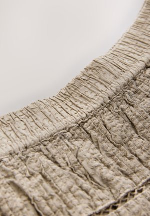 Bež teksturirana tkanina s istaknutim naborima i skupljenim rubom. Površina prikazuje suptilan, nepravilni uzorak za dodatni detalj.