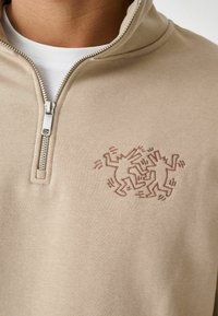 Beige sweatshir med lynlås og høj krave, med et subtilt, mørk beige broderet design af to dansende figurer på venstre bryst.