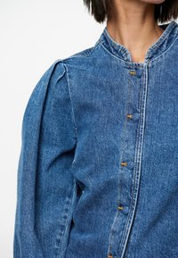 Spijkerjasje met opgeblazen schouders, een hoge halslijn en zichtbare oranje stiksels langs de knoopsluitingen. Klassieke blauwe kleur met een gestructureerde afwerking.