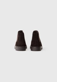 Une paire de bottes Chelsea en suède marron foncé avec des panneaux élastiques noirs sur les côtés et des semelles noires, présentée de dos sur un fond clair.
