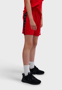 Persoon staand, gekleed in rode sportshorts met zwarte zijstrepen, witte sokken en zwarte sportschoenen tegen een effen achtergrond.