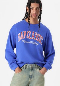 Sudadera de manga larga azul con el atrevido texto "GAP CLASSIC" en rojo y blanco, fabricada en suave tejido y con un corte relajado. Combinada con jeans de lavado claro.