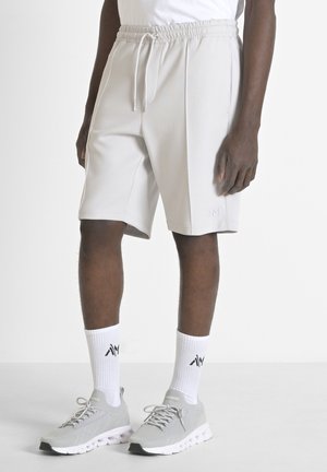 Pantalones cortos deportivos de color gris claro con cintura elástica y bolsillos laterales. Tejido texturizado con un logo sutil en el dobladillo. Llevados con zapatillas grises y calcetines blancos.