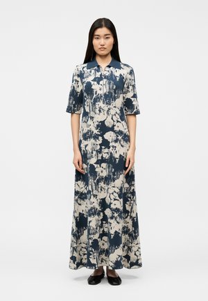 Kobieta stojąca w długiej sukience z krótkim rękawem w granatowo-biały, abstrakcyjny kwiatowy wzór, z ciemnym kołnierzem, w połączeniu z czarnymi balerinami.