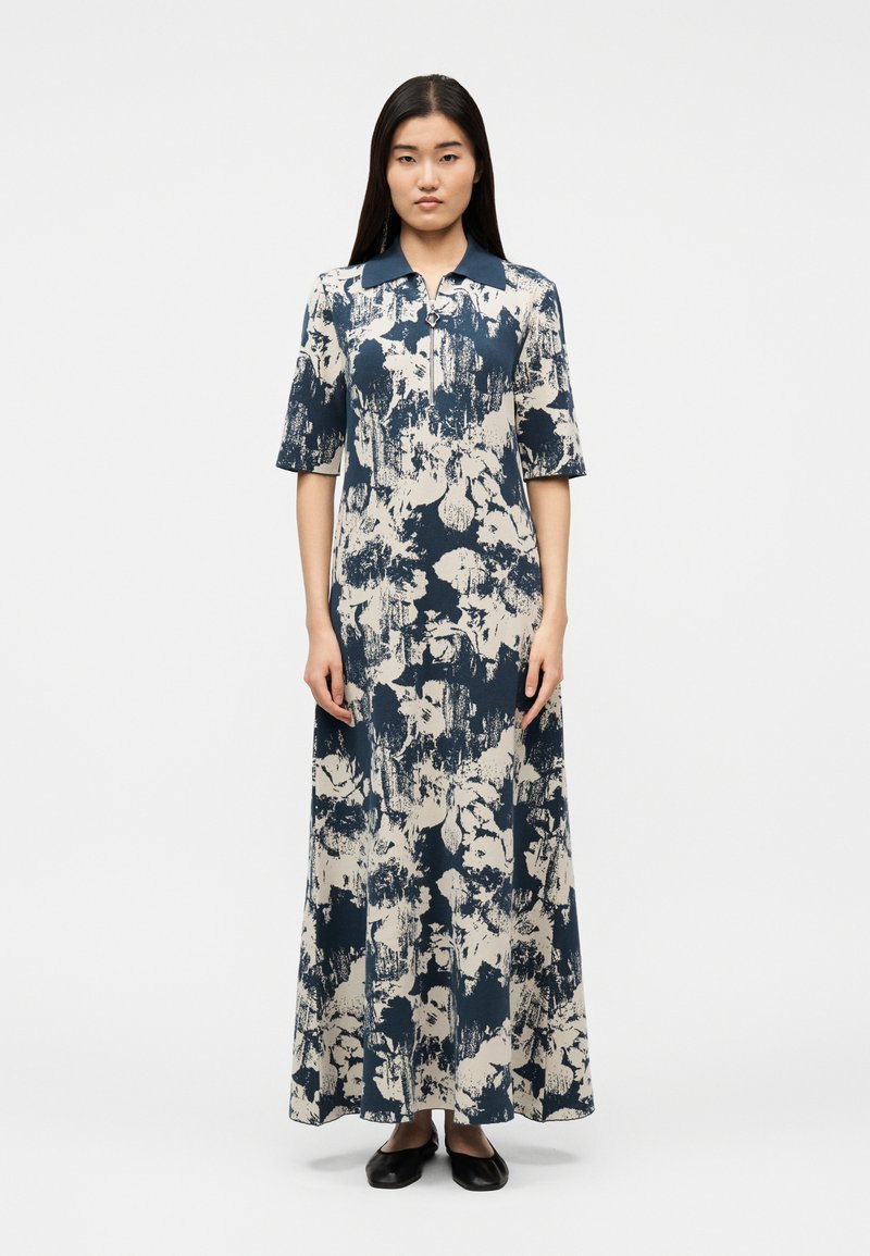 Γυναίκα που στέκεται, φορόντας μακρύ φόρεμα με κοντά μανίκια, σε σχέδιο αφηρημένα λουλούδια σε ναυτικό μπλε και λευκό χρώμα, με σκούρο γιακά, συνδυασμένο με μαύρα φλατ παπούτσια.