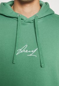 Primer plano de una persona que lleva una sudadera verde con cordones y un logo bordado en estilo de firma blanca en el pecho.