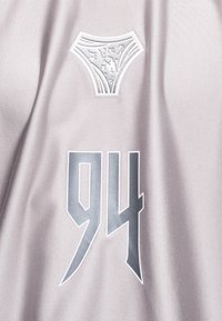 Ασημί ανακλαστικό «94» και ένα στυλιζαρισμένο έμβλημα σε ανοιχτό γκρι υφή ύφασμα, πιθανώς μέρος αθλητικής φανέλας ή ρούχου.