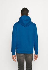 Modrá mikina s kapucí z měkké tkaniny s velkou kapucí, žebrovanými manžetami a lemem. Pohled zezadu ukazuje hladkou texturu a volný střih.