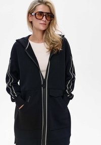 Czarna bluza z kapturem na zamek błyskawiczny, z kontrastującymi białymi szwami wzdłuż rękawów, przednimi kieszeniami i kapturem. Wykonana z miękkiego, teksturowanego materiału.