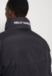 Zwarte pufferjack met hoge kraag. "HELly HANSEN" logo gedrukt in het wit op de achterkant. Gladde textuur met gestikte paneeldetails.