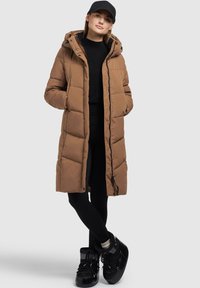 Brauner gepolsterter langer Mantel mit Kapuze, ausgestattet mit einem Reißverschluss und Seitentaschen, kombiniert mit schwarzen Leggings und glänzenden schwarzen High-Top-Stiefeln.