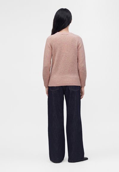 Femme aux longs cheveux noirs portant un pull rose clair et un jean large foncé, debout de dos sur un fond blanc uni.