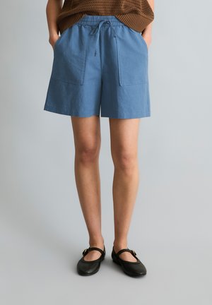 Persoon draagt blauwe shorts met trekkoord en grote voorzakken, bruin gebreid topje en zwarte Mary Jane platte schoenen, staand tegen een grijze achtergrond.