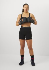 Luvas de boxe pretas com detalhes laranja, combinadas com um top desportivo preto e calções de cintura subida. Footwear são sapatilhas desportivas escuras com detalhes vermelhos.