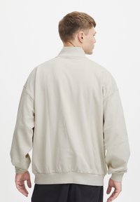 Sudadera de color beige con un cuello alto, costuras de hombro caídas y puños acanalados. Textura suave y ajuste relajado, sin patrones visibles.