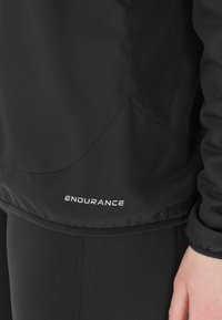 Svart lättviktsjacka med en slät textur, prydd med märket "ENDURANCE" i vitt vid fållen. Jackan har en figursydd design och elastiska ärmslut.