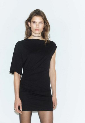 Un mannequin se tient devant un fond pâle, portant une mini-robe noire ajustée avec un col asymétrique et un collier double tour.