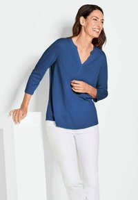 Blaues Langarmshirt mit V-Ausschnitt, abgerundetem Saum und weicher Textur, kombiniert mit weißen Hosen. Das Modell steht vor einem weißen Hintergrund.