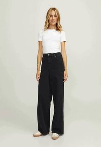 Calças largas pretas com cintura alta, fecho de botão e grandes bolsos, combinadas com uma t-shirt justa branca e sapatilhas claras.