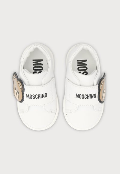 Chaussures blanches pour bébé avec logo Moschino sur les brides et détail visage d’ourson sur les côtés extérieurs, vues de dessus sur fond clair.