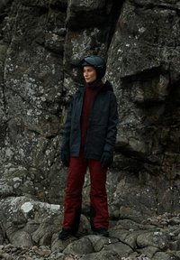 Svart vattentät jacka, mörkröd fleece, och matchande byxor, med svarta handskar och hjälm, stående på stenig terräng. Grå, texturerad bakgrund.