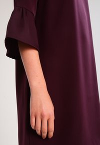 Arm och hand på en person klädd i en långärmad, mörklila klänning med ett löst, utsvängt manschettdesign.