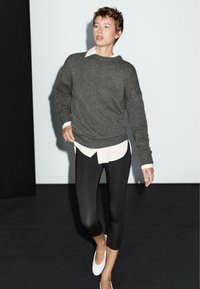 Grå stickad tröja över en vit skjortkrage, kombinerad med svarta leggings och vita skor, på en mörk bakgrund.