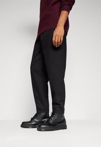 Henkilö, jolla on päällään mustat nauhakengät ja löysät mustat housut, sekä viininpunainen tekstiilillä varustettu neule, seisoo tasaista taustaa vasten.