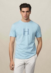 T-shirt en coton bleu clair avec un grand graphique "H" et le texte "Hackett" en dessous, col rond et manches courtes.