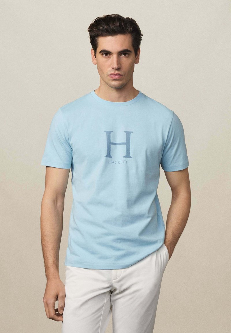 T-shirt en coton bleu clair avec un grand graphique "H" et le texte "Hackett" en dessous, col rond et manches courtes.