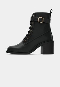 Bottine en cuir noir avec lacets sur le devant, talon épais, bride latérale avec boucle dorée et languette à l'arrière.