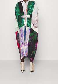 Robe longue multicolore avec divers motifs abstraits, présentant un tissu plissé et un décolleté en V. Accompagnée de bottines beige clair à la cheville.