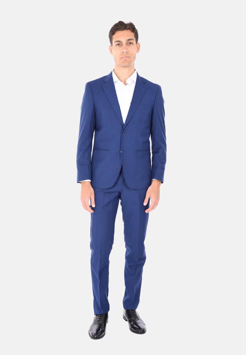 Completo blu navy con un design aderente, giacca monopetto e pantaloni dritti. Camicia bianca sotto e scarpe nere lucide.