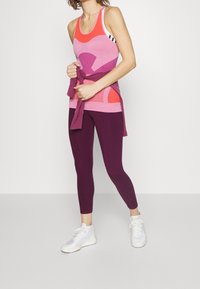 Vaaleanpunainen ja violetti urheilutoppi geometrisilla kuvioilla, yhdistettynä syvän violetit legginsit; malli on sitonut täydentävän topin vyötärölle.