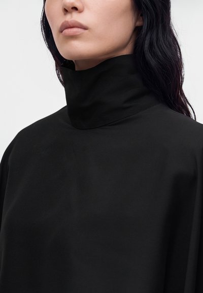 Gros plan d'un mannequin portant un haut noir à col montant sur un fond blanc uni.