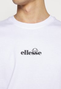 Gros plan d'une personne portant un t-shirt blanc avec un petit logo noire ellesse sur la poitrine sous l'encolure.