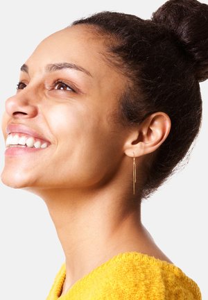 Goldene, hängende Ohrringe mit dünnem, elongated Design. Das Model hat einen glatten Teint und trägt einen gelben, strukturierten Pullover. Schlichter und eleganter Stil.