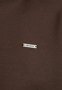 Brauner Stoff mit einer glatten Textur und einem silbernen Metalltag, auf dem "SMILODOX" steht, der mit zwei Löchern zum Befestigen versehen ist.