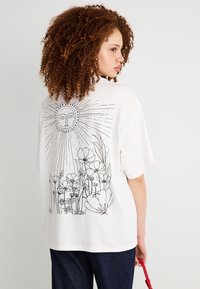 Γυναίκα με σγουρά μαλλιά, φορώντας ένα λευκό φαρδύ T-shirt με μαύρα γραμμικά σχέδια ενός ήλιου και λουλουδιών στην πλάτη, κρατώντας μια κόκκινη τσάντα.