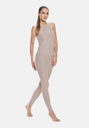 Hosszú hajú nő bézs színű ujjatlan felsőben és leggingsben, mezítláb áll, egyik lábát előre helyezve, egyszerű háttér előtt.