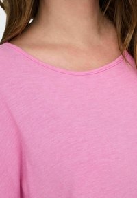 T-shirt rosa chiaro con un'ampia scollatura rotonda, maniche corte e tessuto morbido, leggermente texturizzato. Dettagli delle cuciture sulle spalle.
