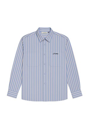 Junger Mann mit cremefarbenem Baseball-Cap, blau-weiß gestreiftem Hemd über einem weißen T-Shirt mit Logo, dunkelgrüne Hose, neutraler Gesichtsausdruck.