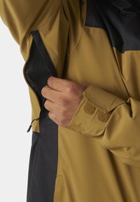 Veste résistante à l'eau présentant un design en coloris marron et noir, une poche en mesh et des poignets en Velcro ; texture lisse et matériau durable.