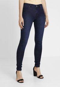 Kvinna iklädd mörkblå tajta jeans och svarta högklackade sandaler, som står mot en enkel ljus bakgrund.