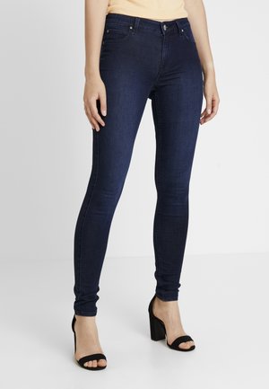 Femme portant un jean skinny bleu foncé et des sandales à talons noirs, debout devant un fond clair et uni.