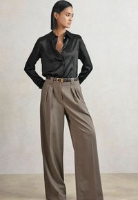 Schwarze Satin-Hemdenjacke mit leicht geöffnetem Kragen, kombiniert mit hoch taillierten, gefalteten beigen Hosen und einem braunen gemusterten Gürtel.
